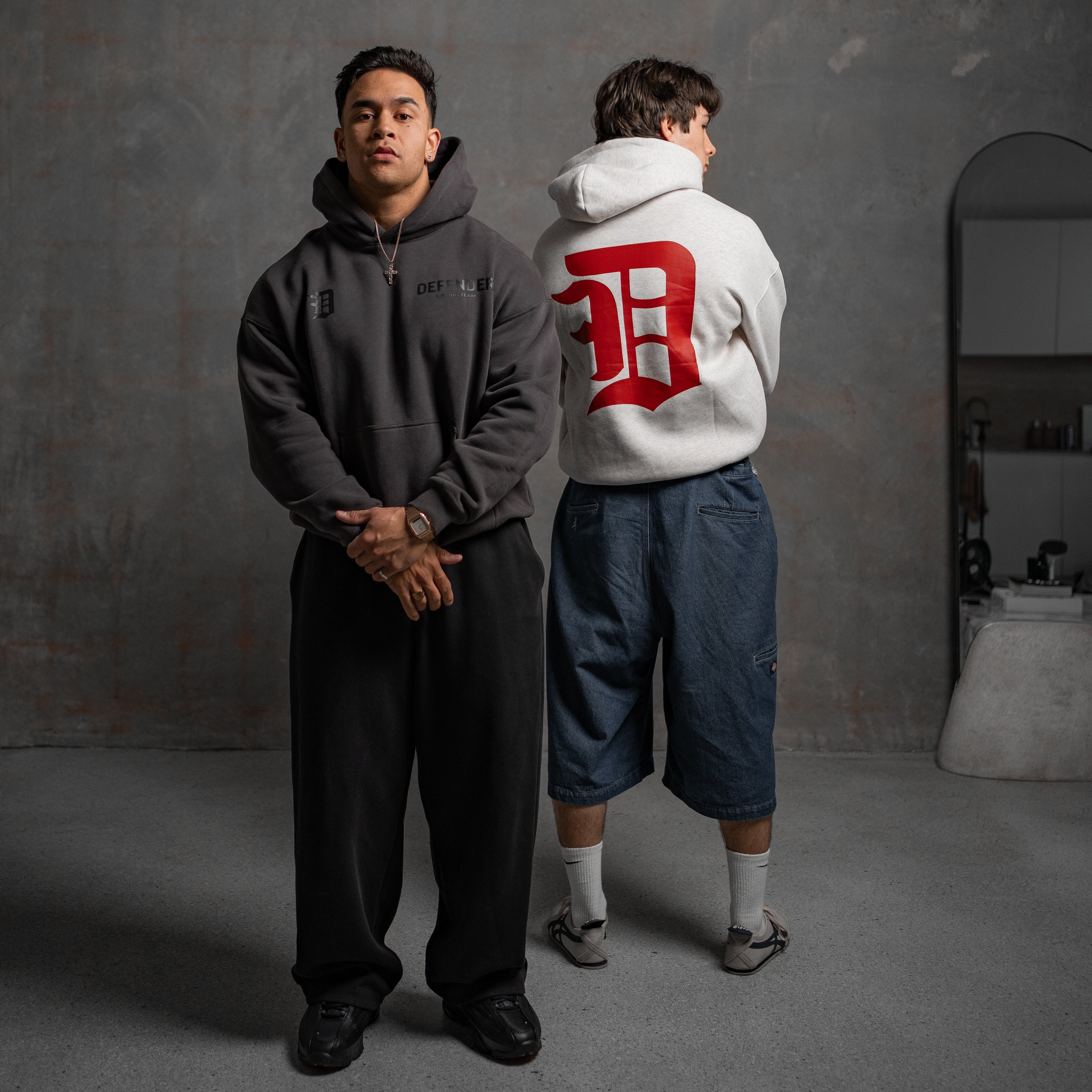 Two individuals wearing hoodies in a room with a gray wall.